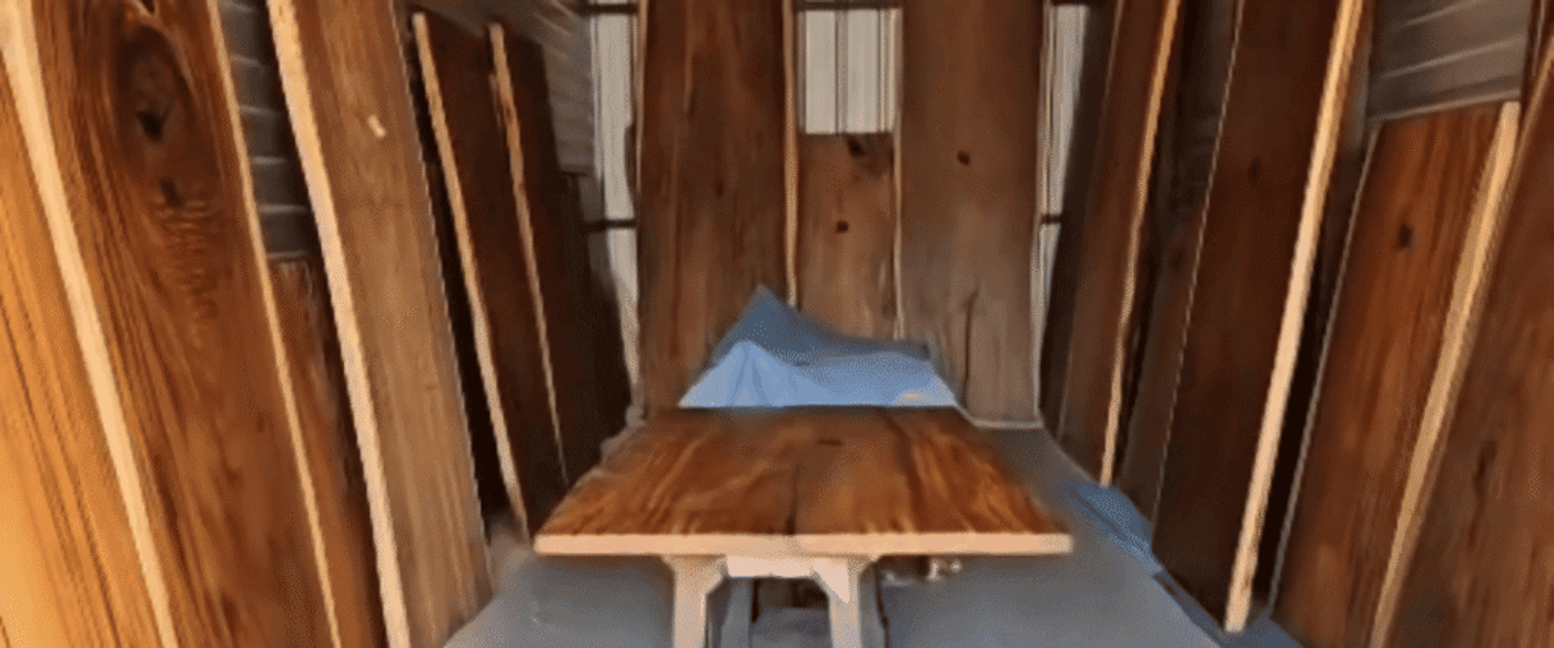 Hardwood planks stacked vertically and a finished wooden tabletop on sawhorses inside a workshop, warm wood grain visible.