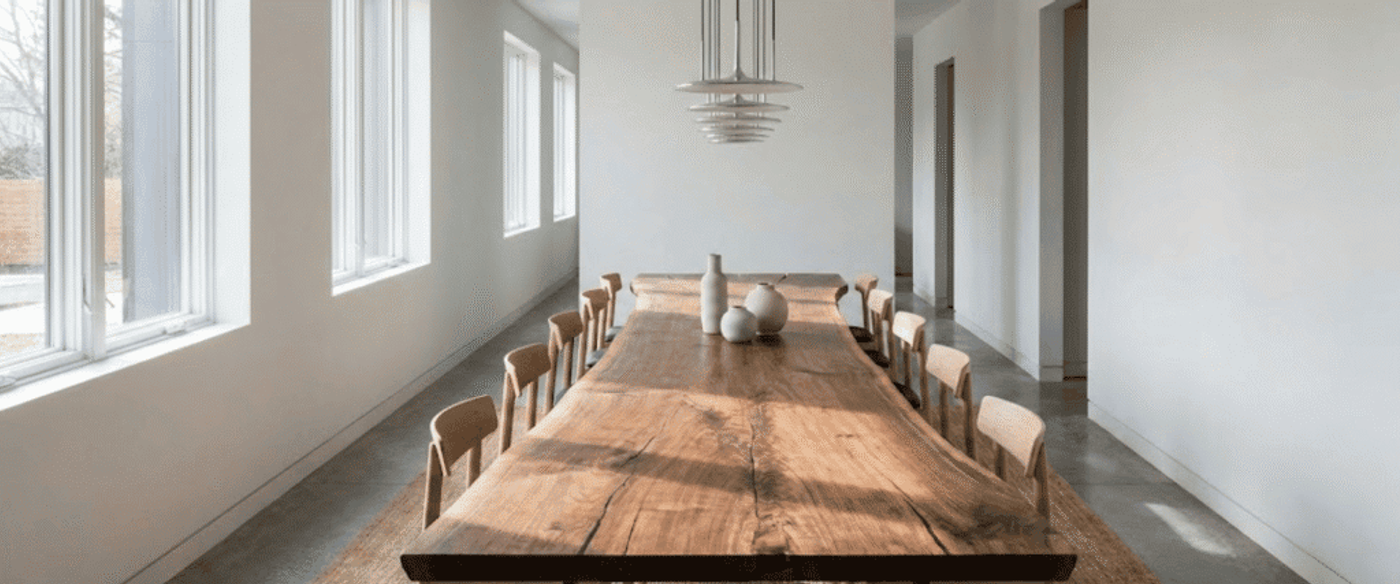 Long natural wood dining table with chairs in a bright minimalist room, sunlight streaming through tall windows, simple pendant light