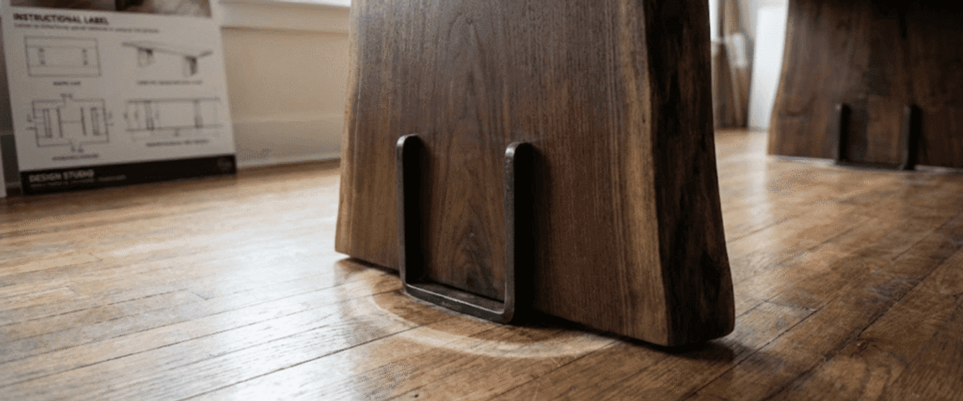 Solid wood table base with metal U-bracket support on worn hardwood floor, soft daylight from window and assembly diagram in background