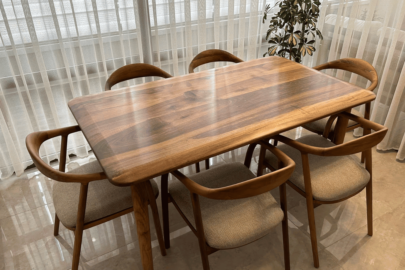 Modern wooden dining table with natural finish and six cushioned chairs in a bright room with sheer curtains and plant