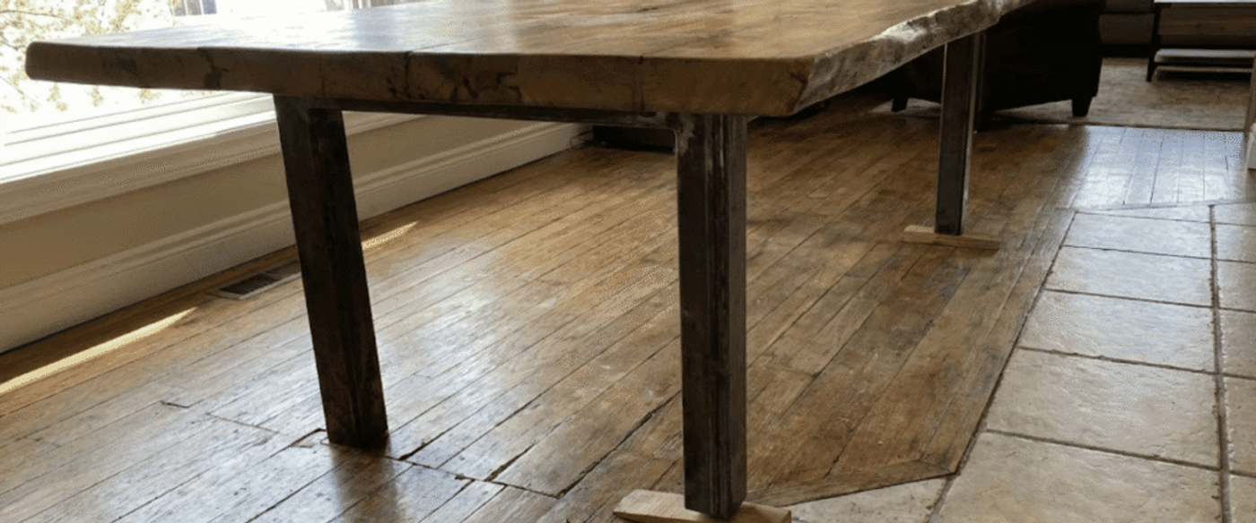 Live-edge wooden dining table with metal legs on reclaimed hardwood and tile floor in a sunlit living room