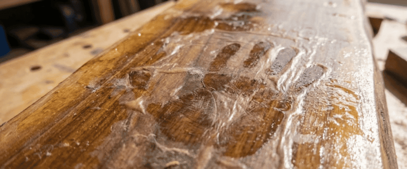 Handprint pressed into wet wood finish on a workbench, glossy grain and sawdust visible across the oak plank