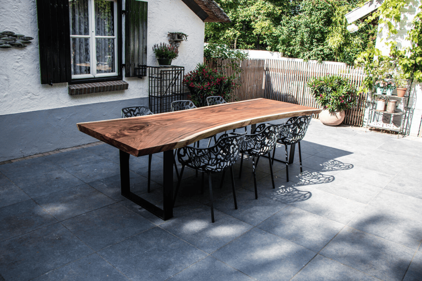 Outdoor wooden dining table with black metal legs and six black patterned chairs on a tiled patio next to a house with white walls, black shutters, and potted plants.