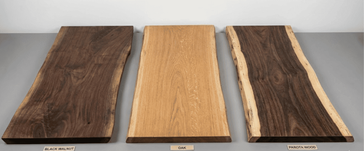 Three long wood slabs on a gray surface: dark black walnut (left), light oak (center), and live-edge parota wood