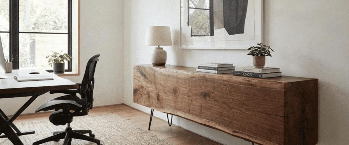 Home office with wooden desk and ergonomic chair by window, live-edge wooden console with lamp, books and potted plants
