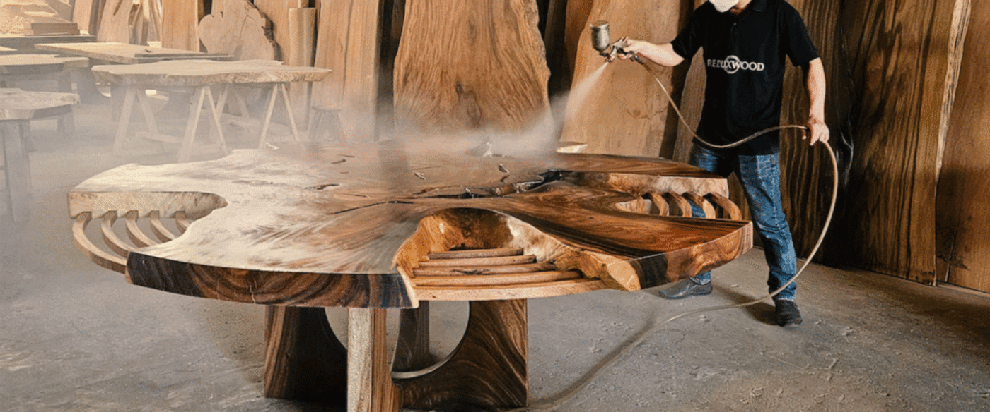 Man spraying finish on a large wooden table in a workshop, enhancing its natural grain and texture.