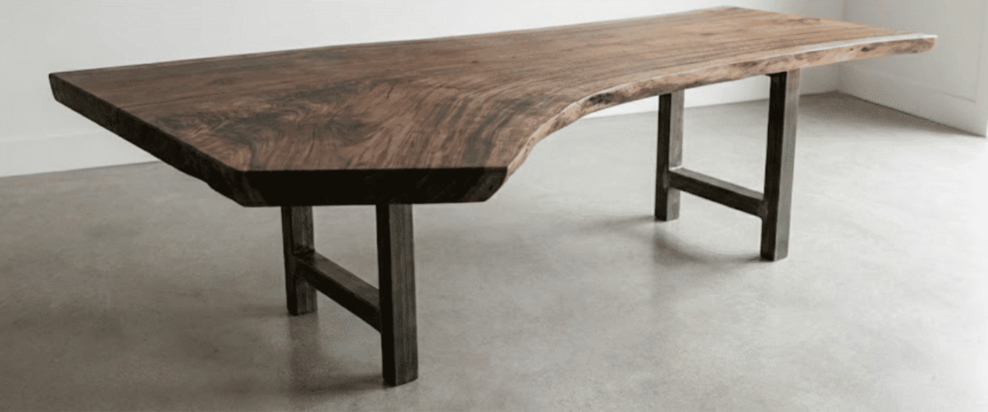 A live-edge wooden table with asymmetrical weight distribution, thick slab top and metal legs set on a concrete floor near a bright window