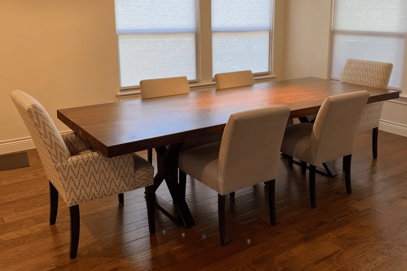 Dining room with a long wooden table and six mixed-pattern upholstered chairs on hardwood floors under three shaded windows.