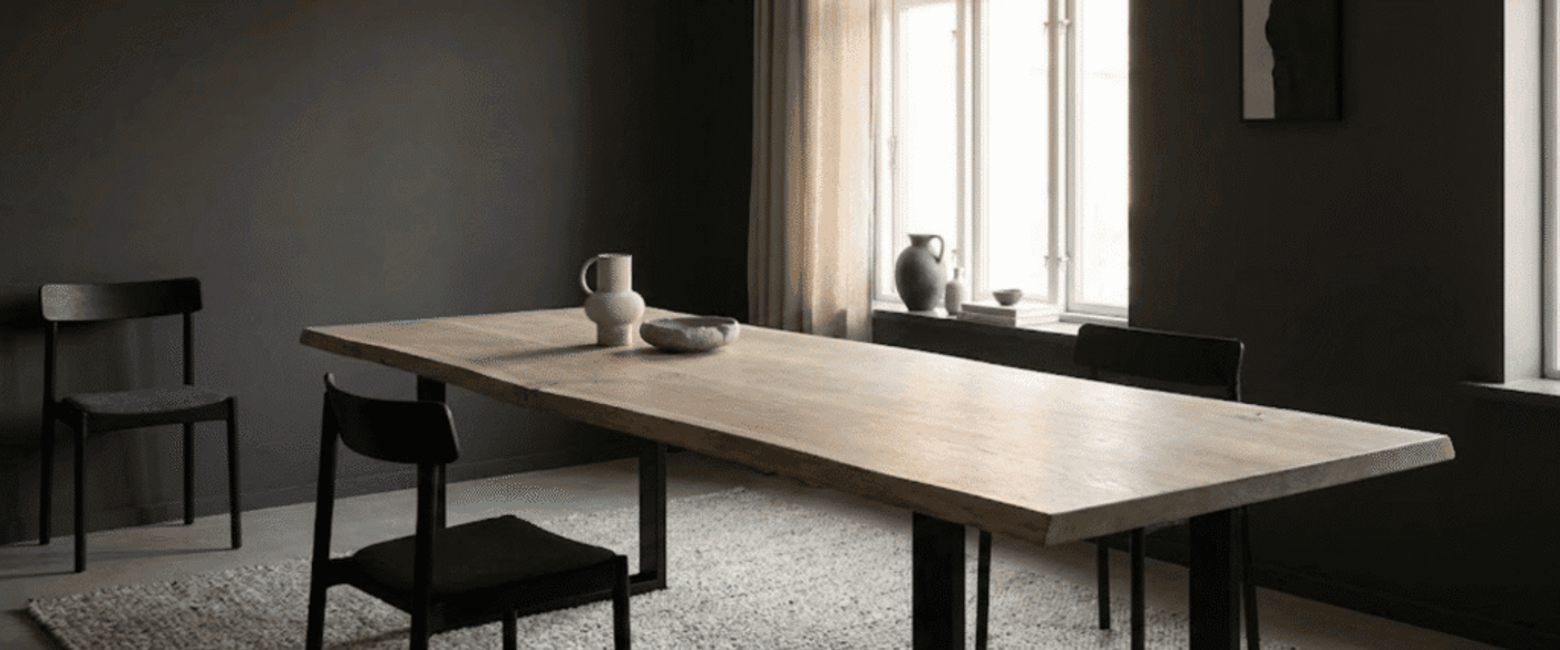 Minimal dining room with a long wooden table, black chairs, neutral pottery on table and windows letting in soft daylight.