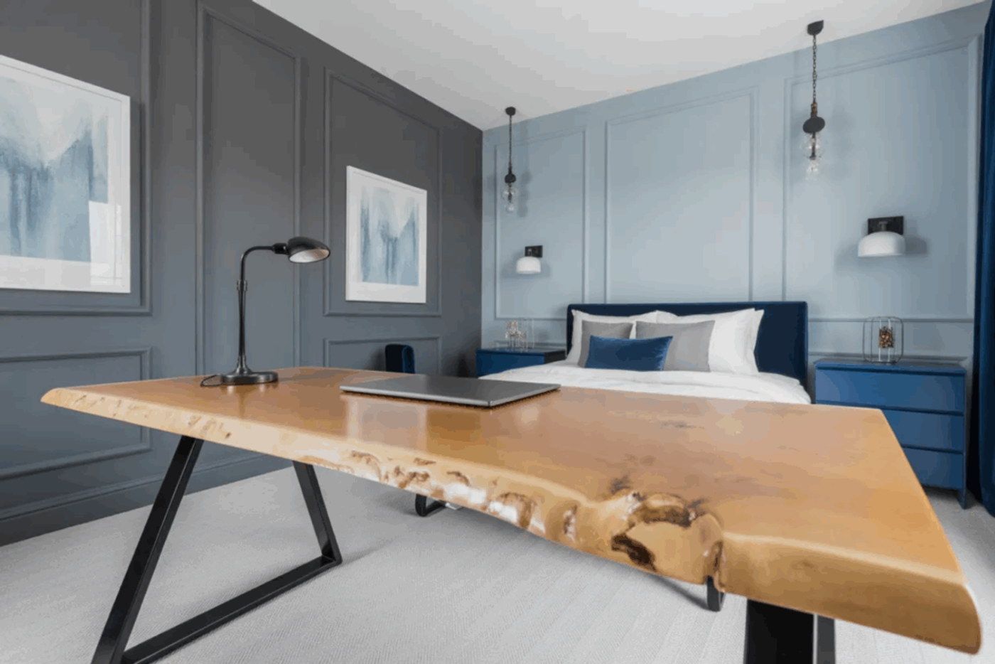 Live edge wooden desk with black metal legs in a modern bedroom featuring blue and gray walls, a bed with blue and white bedding, and minimalist decor.