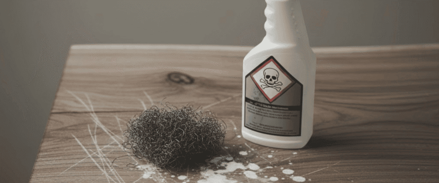 White spray bottle with skull-and-crossbones hazard label beside a steel wool pad and white chemical splatter on a scratched wooden table