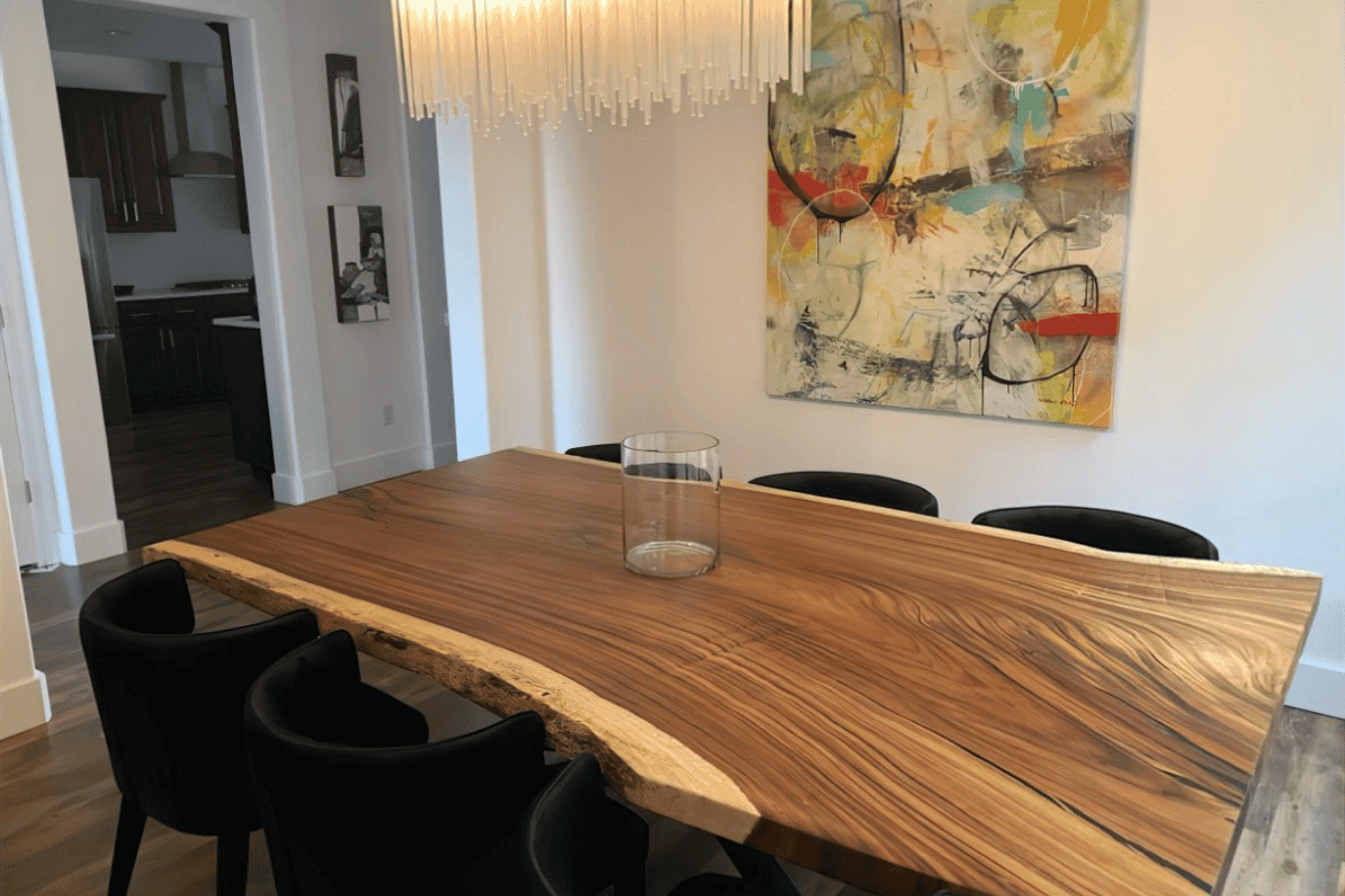 Live-edge walnut dining table with black chairs, glass centerpiece, modern chandelier, and abstract wall painting in bright room.