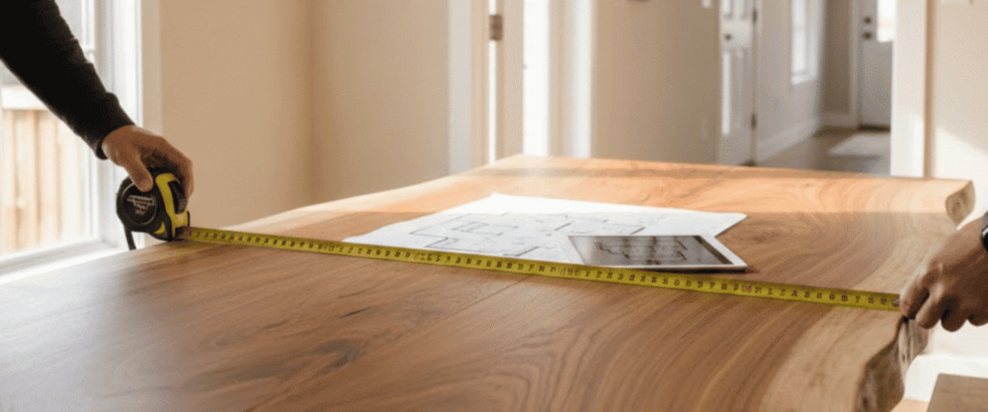 Two people measuring a live-edge wooden table with a tape measure while floor plans rest on the tabletop