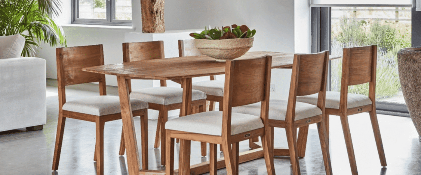 Modern wooden dining table with six cushioned chairs in a bright room, potted centerpiece, large windows and indoor plants.