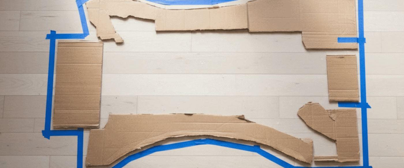 Cardboard template and blue tape outlining a furniture layout on light hardwood floor, with marker and measuring tape nearby