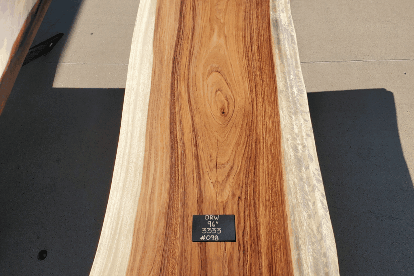 A close-up view of a polished wooden slab with natural wood grain patterns, placed on a concrete surface. The slab has light-colored edges and a rich reddish-brown center. A small black label on the wood shows the text "DRW 96" 3333 #098.
