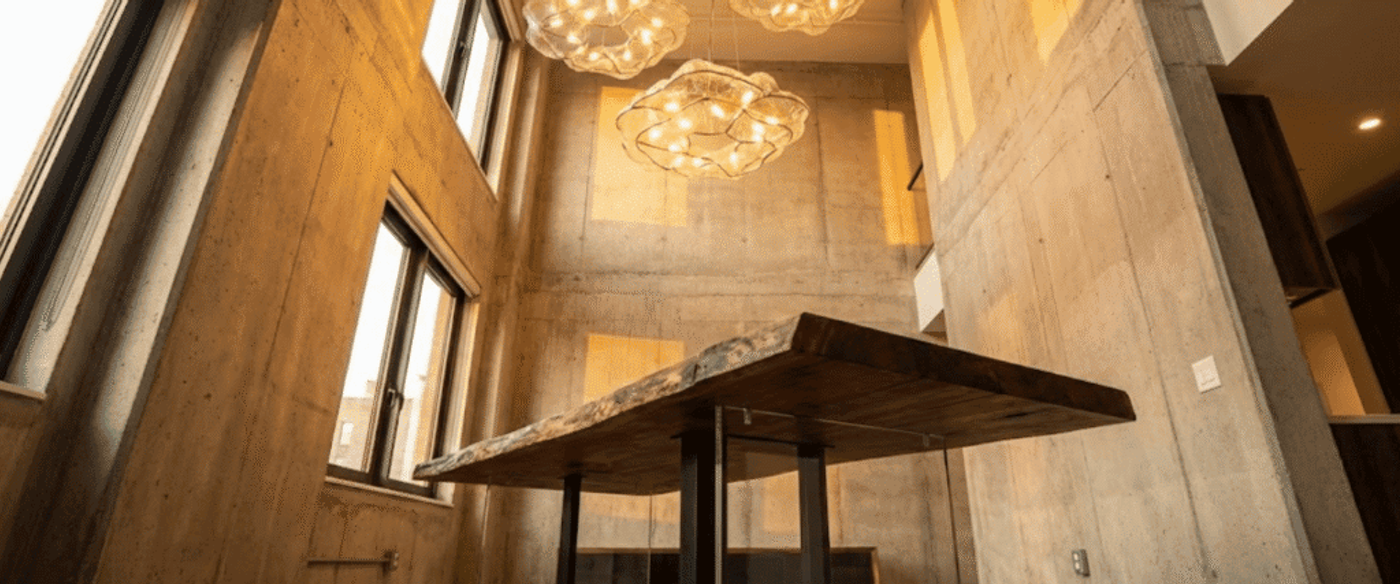 Warm loft dining space with a live-edge wooden table, industrial concrete walls, tall windows, and three glowing cloud-like pendant lights