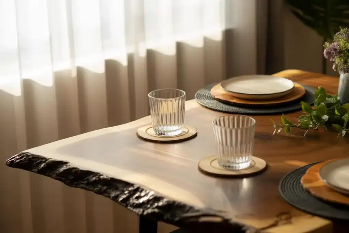 A cozy dining table setup with natural wood texture, two empty glasses on coasters, and neatly arranged plates with placemats, soft sunlight filtering through sheer curtains.