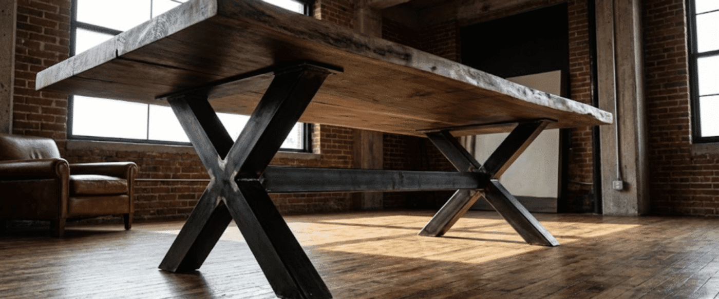 Rustic live-edge wooden table with black metal X-legs in a sunlit industrial loft with brick walls and leather chair