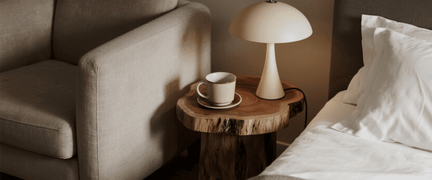 Cozy bedroom corner with a wooden side table, modern lamp, beige armchair, and a cup on a saucer next to a neatly made bed.