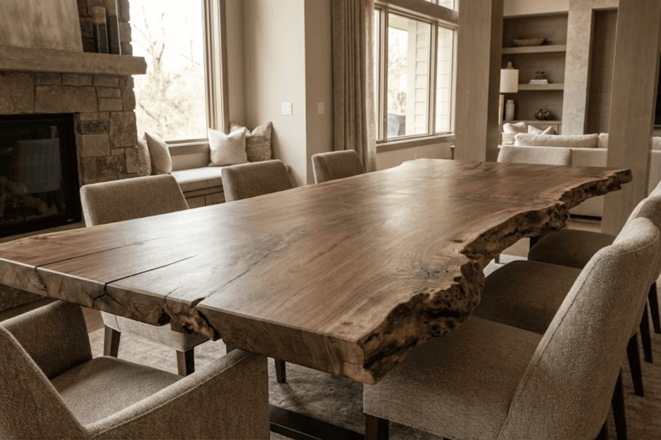 Live-edge walnut dining table surrounded by upholstered chairs in a warm, modern living room with fireplace and large windows