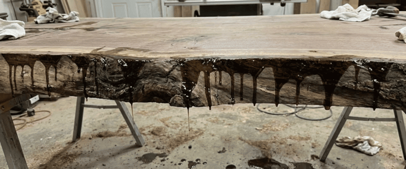 Live-edge walnut slab on sawhorses with dark epoxy resin drips along the rough edge, workshop floor with tools and rags
