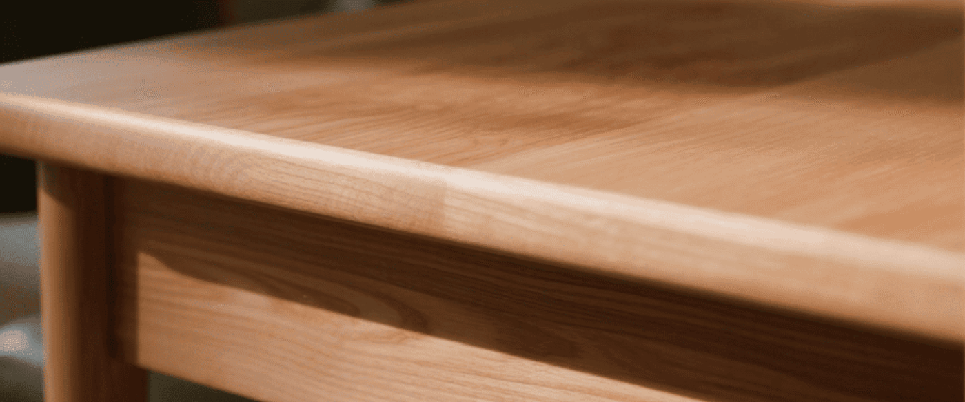 Close-up of a smooth wooden dining table edge and leg with blurred chairs and warm indoor lighting in the background.