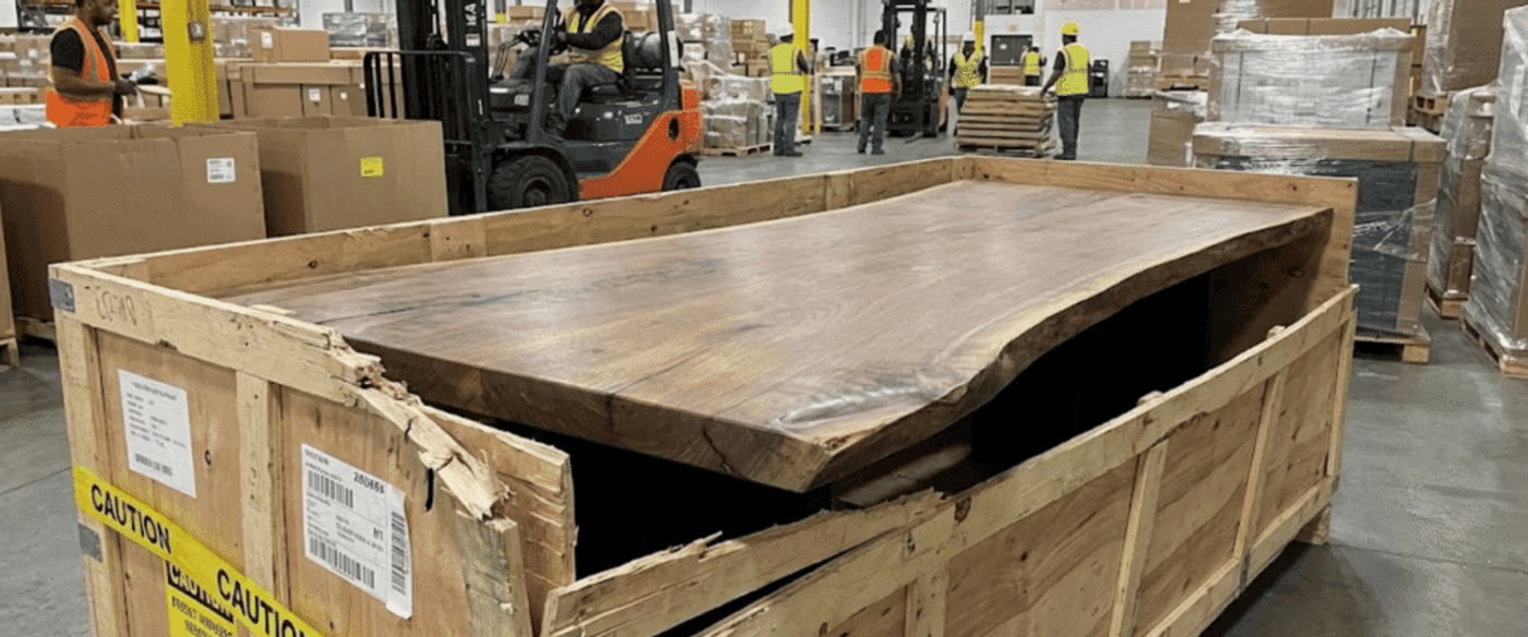 Worker-packed wooden slab table partly exposed in a warehouse crate, damaged crate edge, forklifts and workers in background