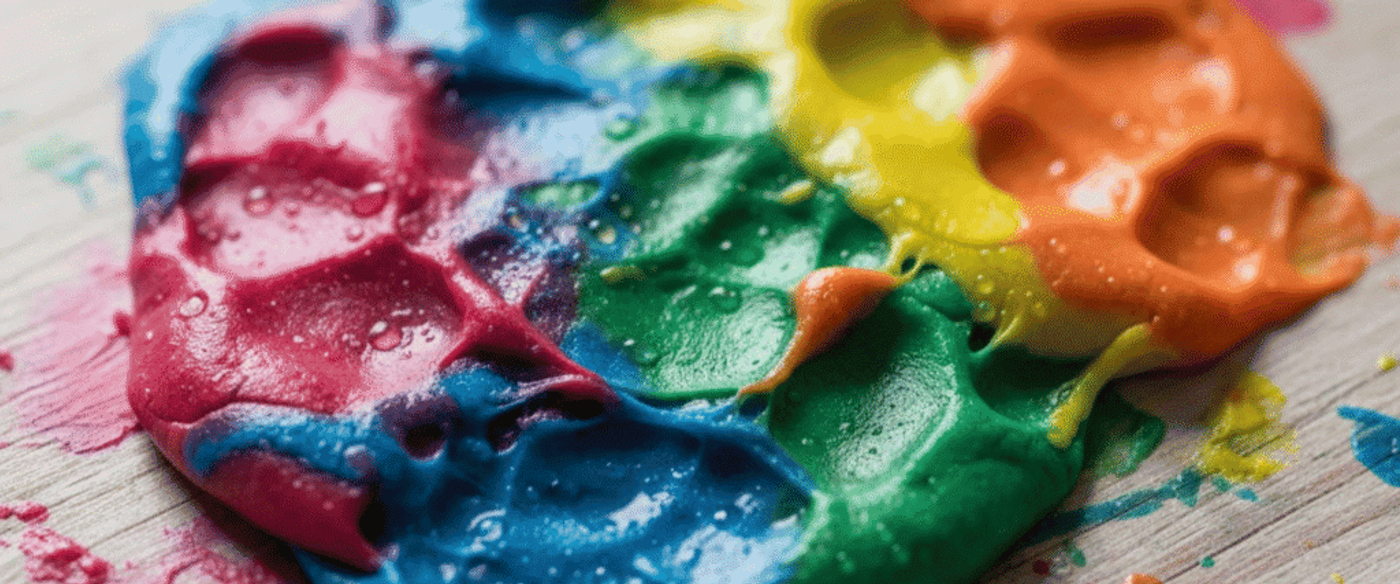 Bright rainbow-colored slime spread on a wooden surface with water droplets and paint smudges, glossy textured puddle