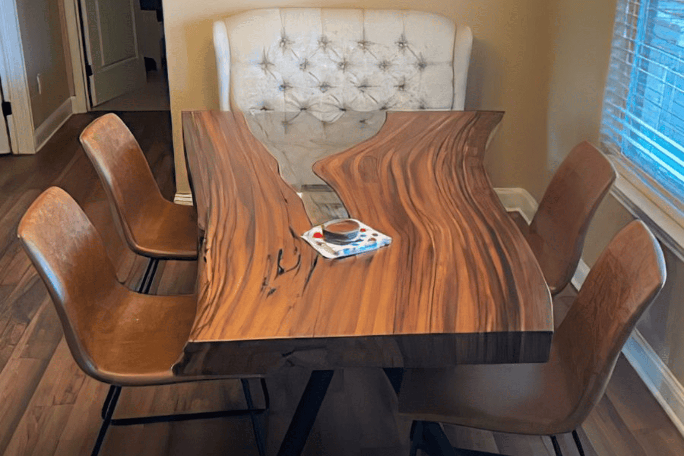 Wooden dining table with natural live edge design, paired with four brown leather chairs and one tufted bench, set in a cozy room with wooden flooring and window blinds.