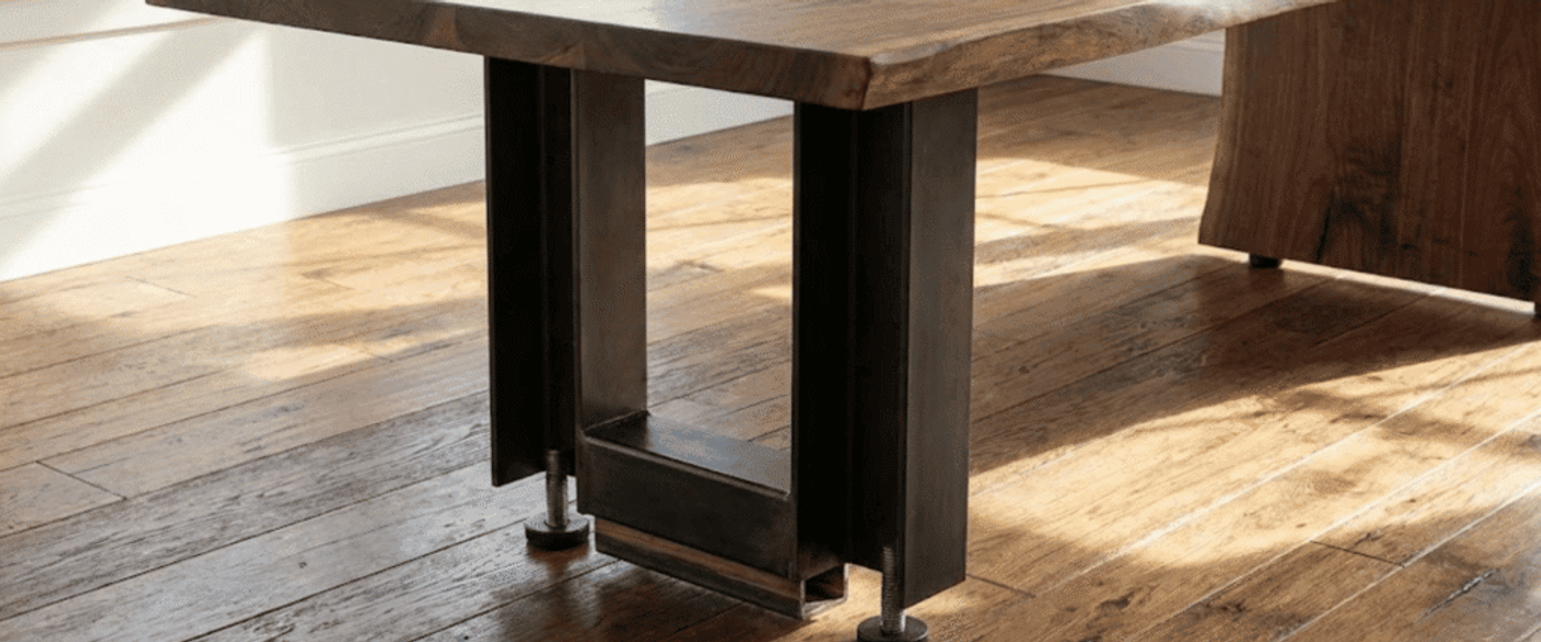 Solid live-edge wooden table top with heavy metal U-shaped leg and adjustable foot on sunlit hardwood floor by a window