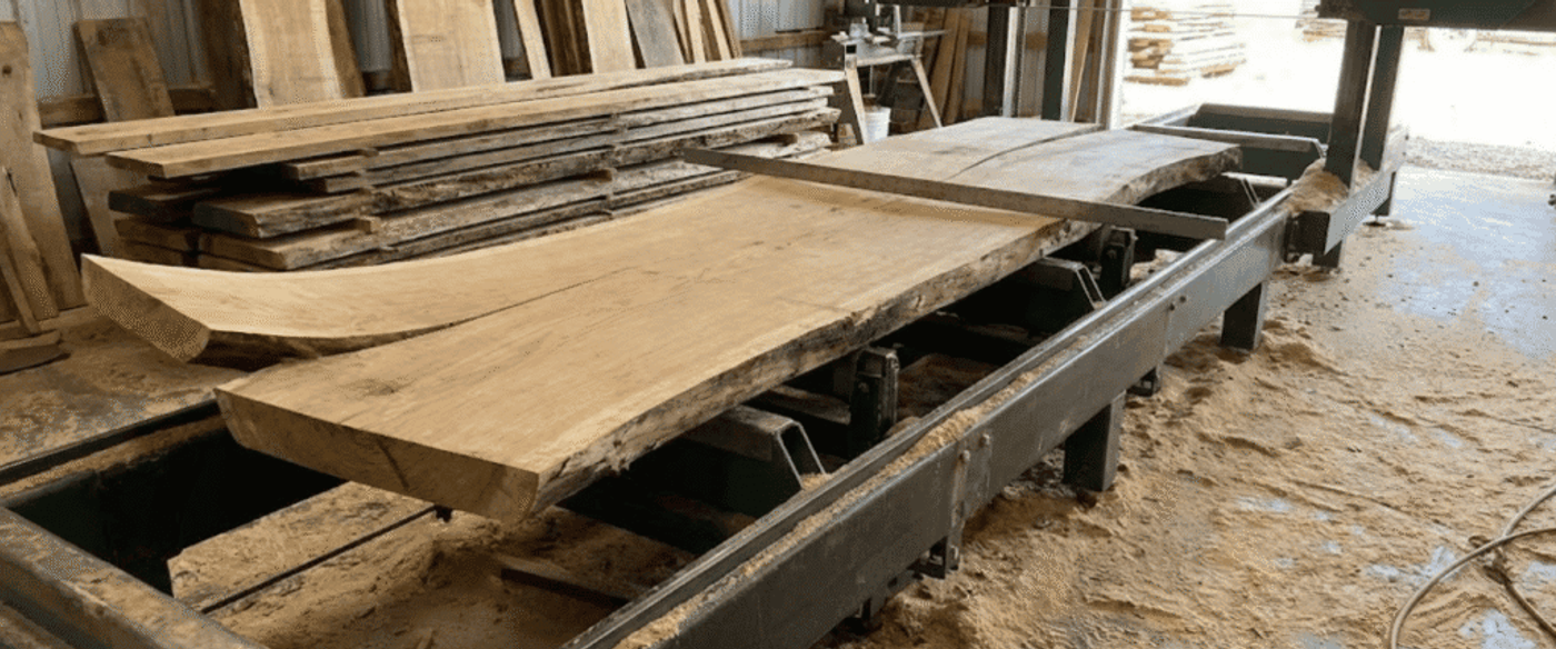 Live-edge oak slabs on a sawmill carriage being cut and stacked inside a workshop, surrounded by lumber and sawdust