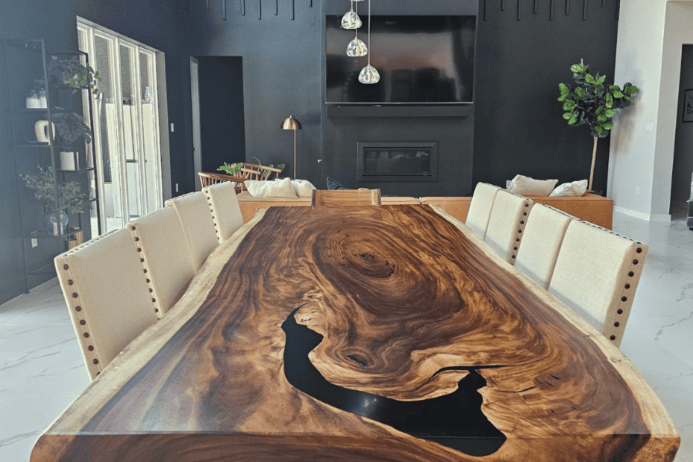 Dining room with a large live edge wood table featuring a natural dark grain and black resin inlay, surrounded by beige upholstered chairs, set in a modern space with black accent wall, TV, plants, and marble floor.