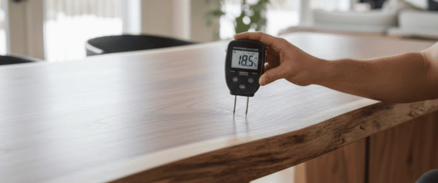 Hand-held moisture meter reading 18.5% inserted into a live-edge wooden table surface in a bright modern dining room