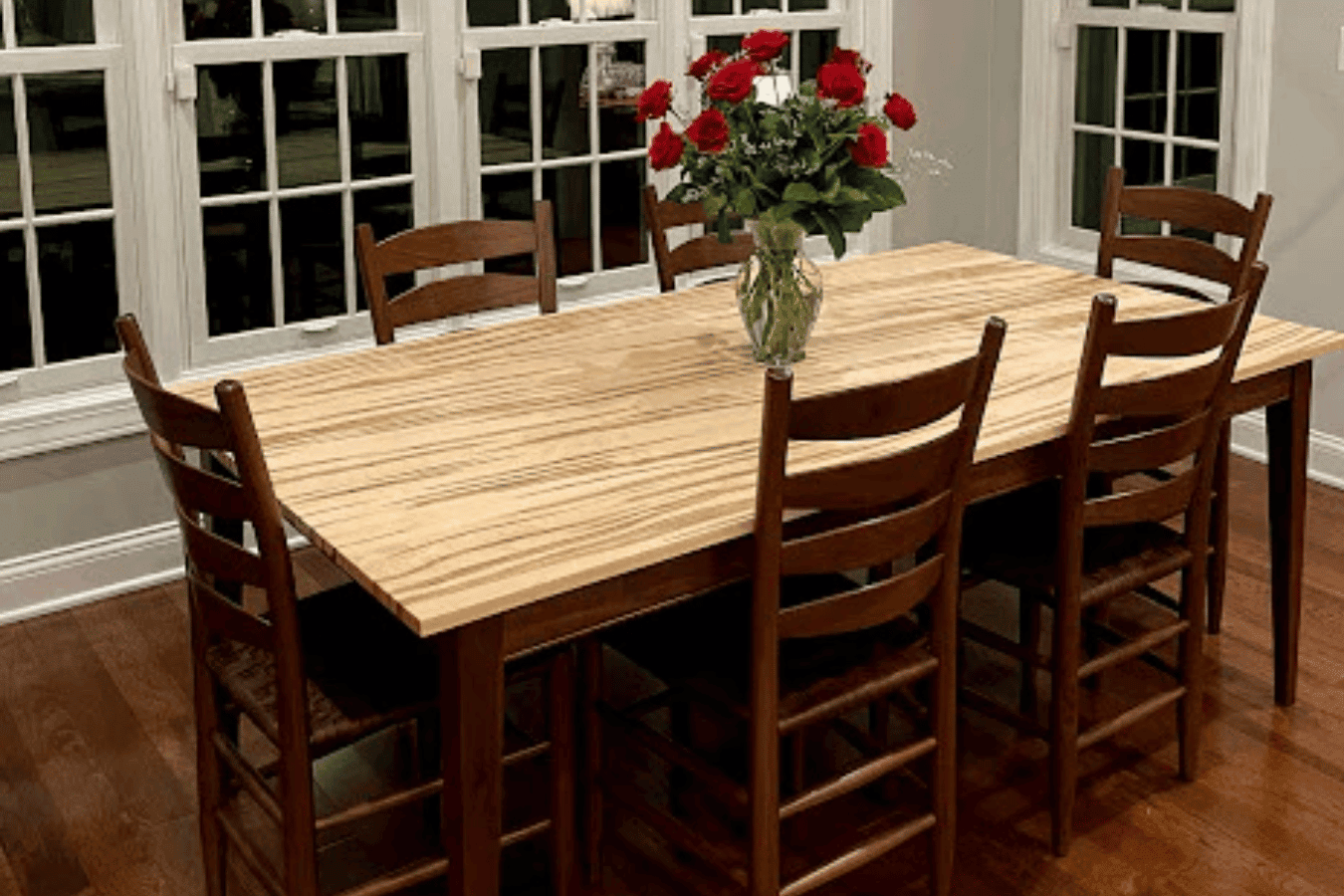 Dining table with six wooden chairs and vase of red roses centered on a light wood table, set beside bright windows.