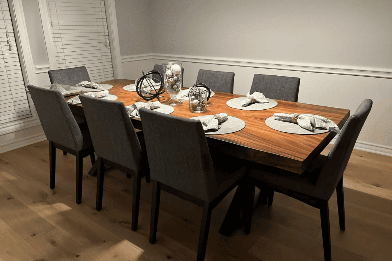 Modern wooden dining table set for eight with gray upholstered chairs, round placemats and folded napkins, decorative centerpiece.