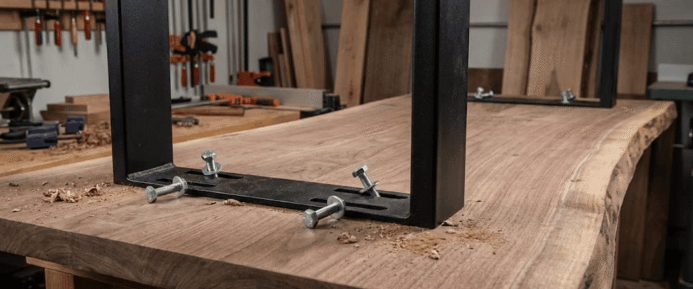 Black metal table legs bolted to a live-edge walnut tabletop in a woodworking shop with clamps