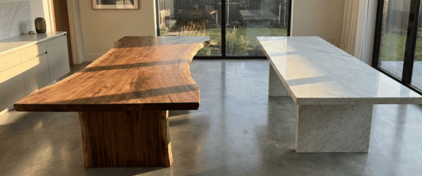 Two large dining tables side-by-side: a live-edge walnut wood table with pedestal base and a sleek white marble table in a sunlit modern room