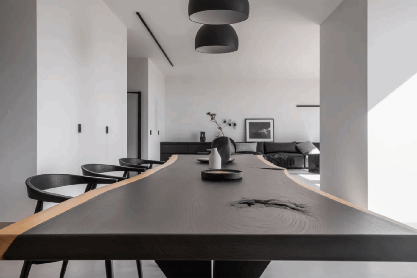 Dark live edge wooden dining table with black chairs in a minimalist white and black modern room with pendant lights and a sofa in the background.