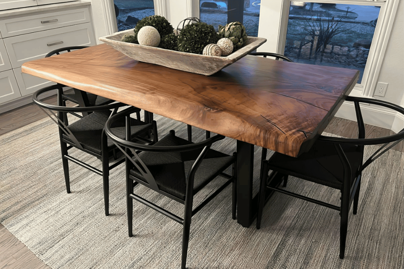 Live-edge wooden dining table with black chairs on a woven rug, centerpiece bowl of greenery and decorative orbs by bay windows.