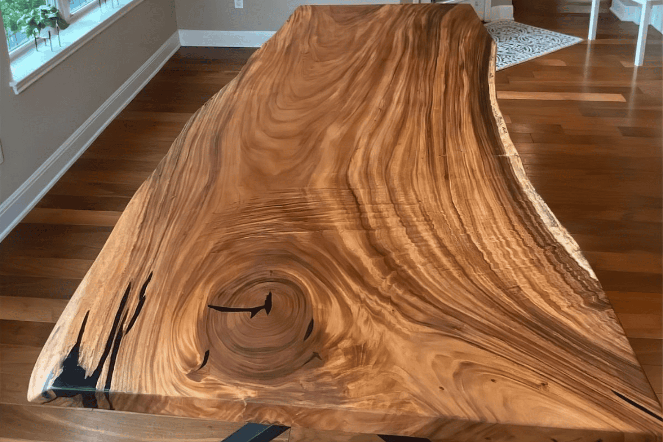Live edge wood table with rich grain patterns and natural knots, placed in bright room with wooden floor.