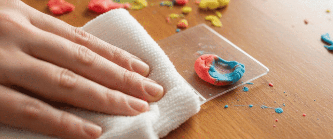 Hand wiping colorful clay pieces and smudges from a wooden table with a white cloth, cleaning up a small craft project mess