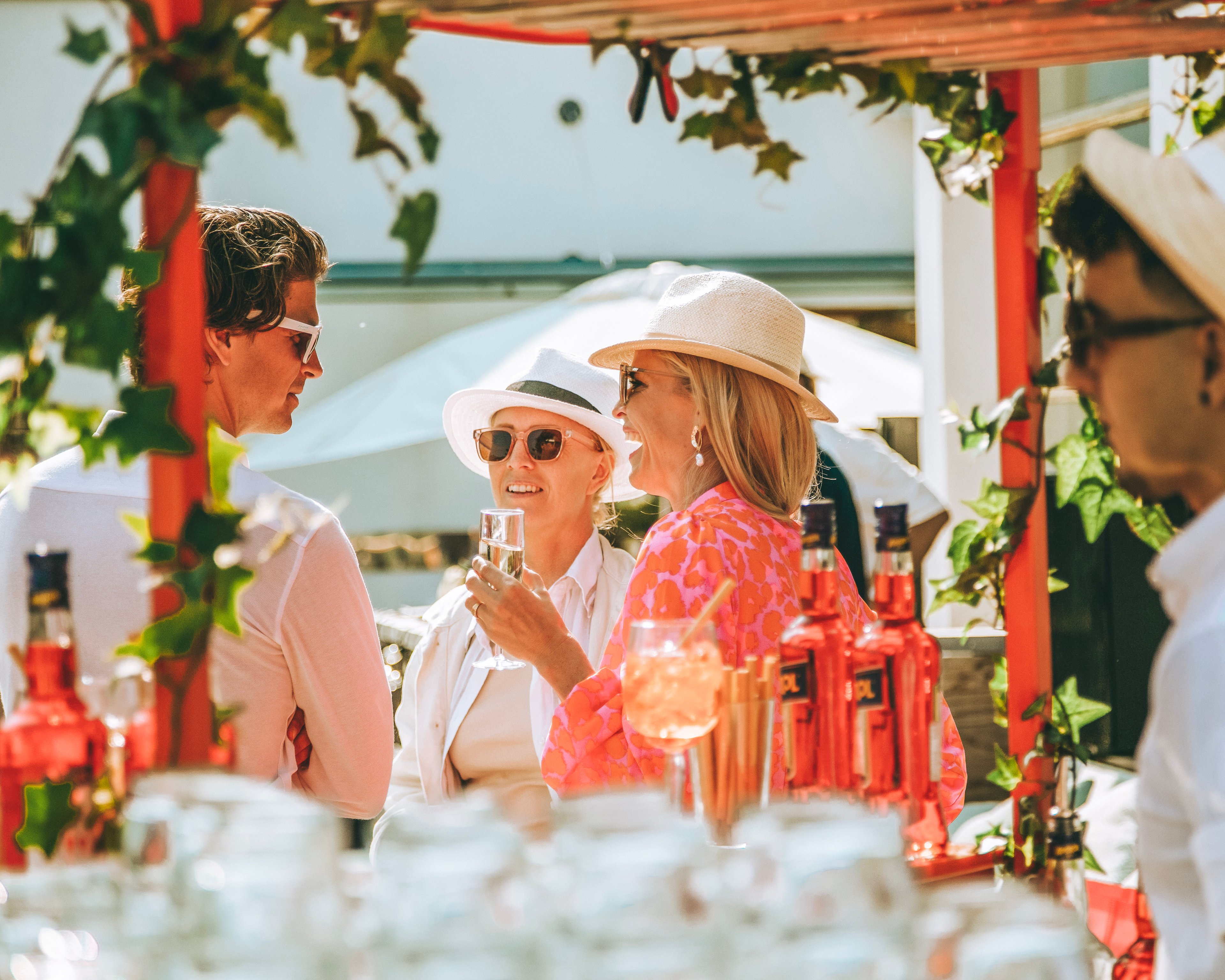 Människor i sommarkläder pratar och skrattar under en murgröneklädd pergola