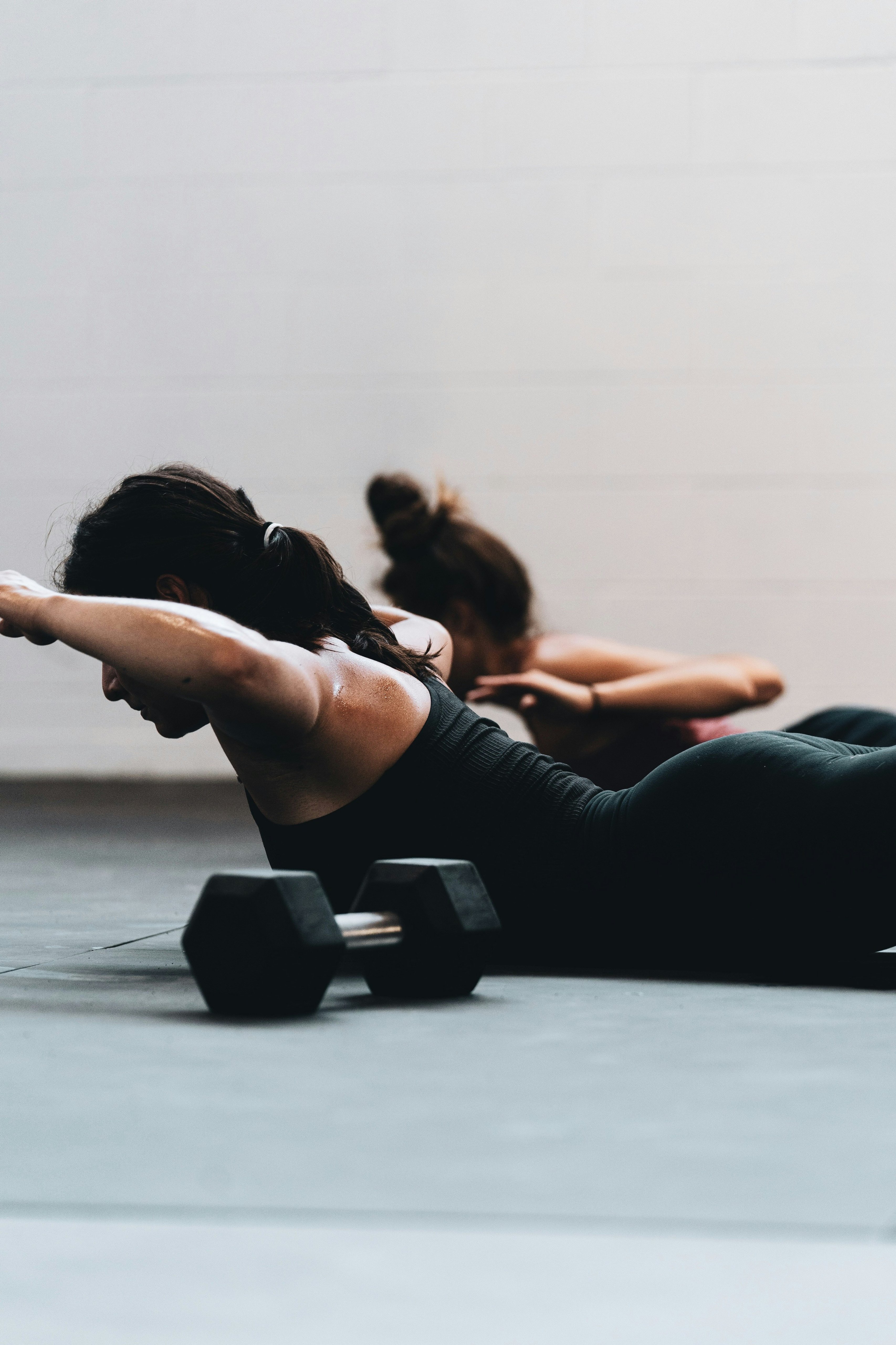 Kvinnor ligger på mage och lyfter armarna i en övning, med en hantel i förgrunden