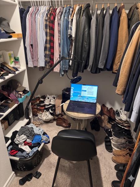 My closet recording studio — a side table, laptop, mic on a boom arm, and a stool surrounded by clothes