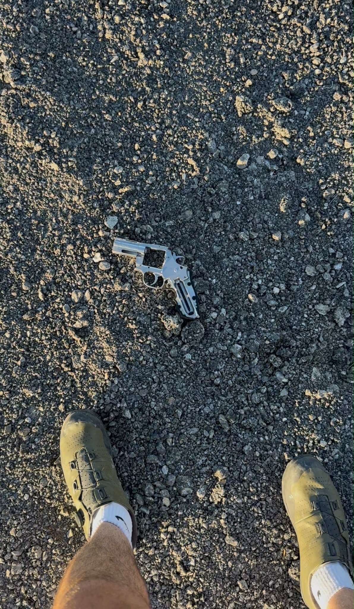 A revolver lying in the gravel of a road shoulder, between two cycling shoes