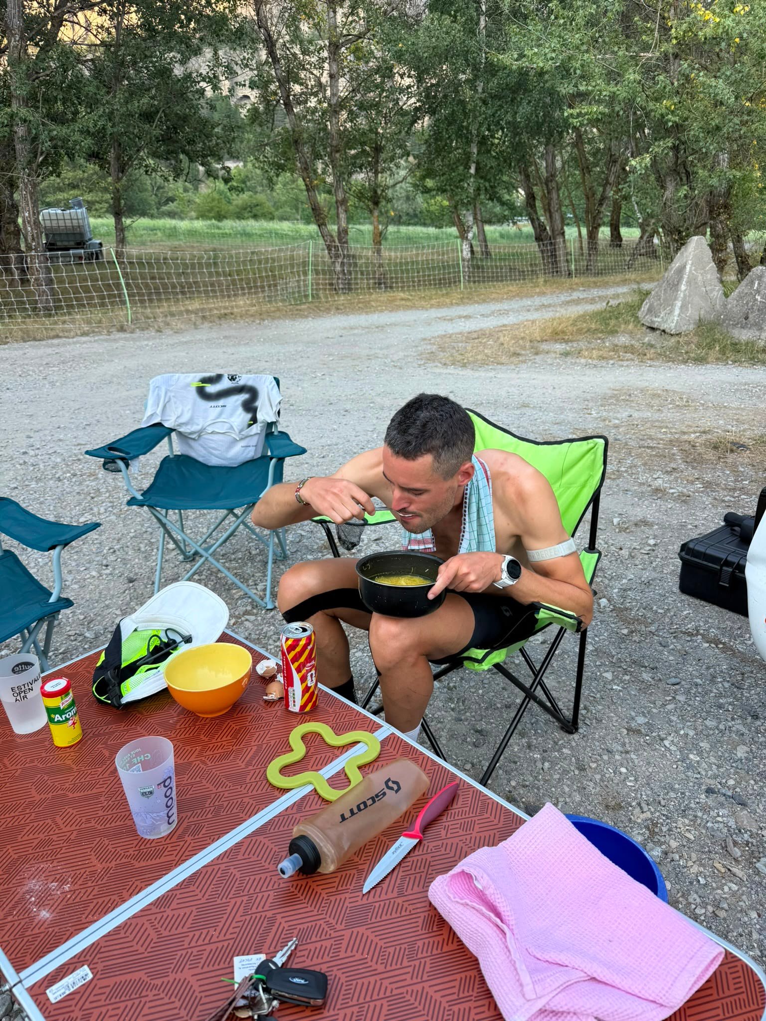 A man eating from a pot in a camp chair beside a camp table.