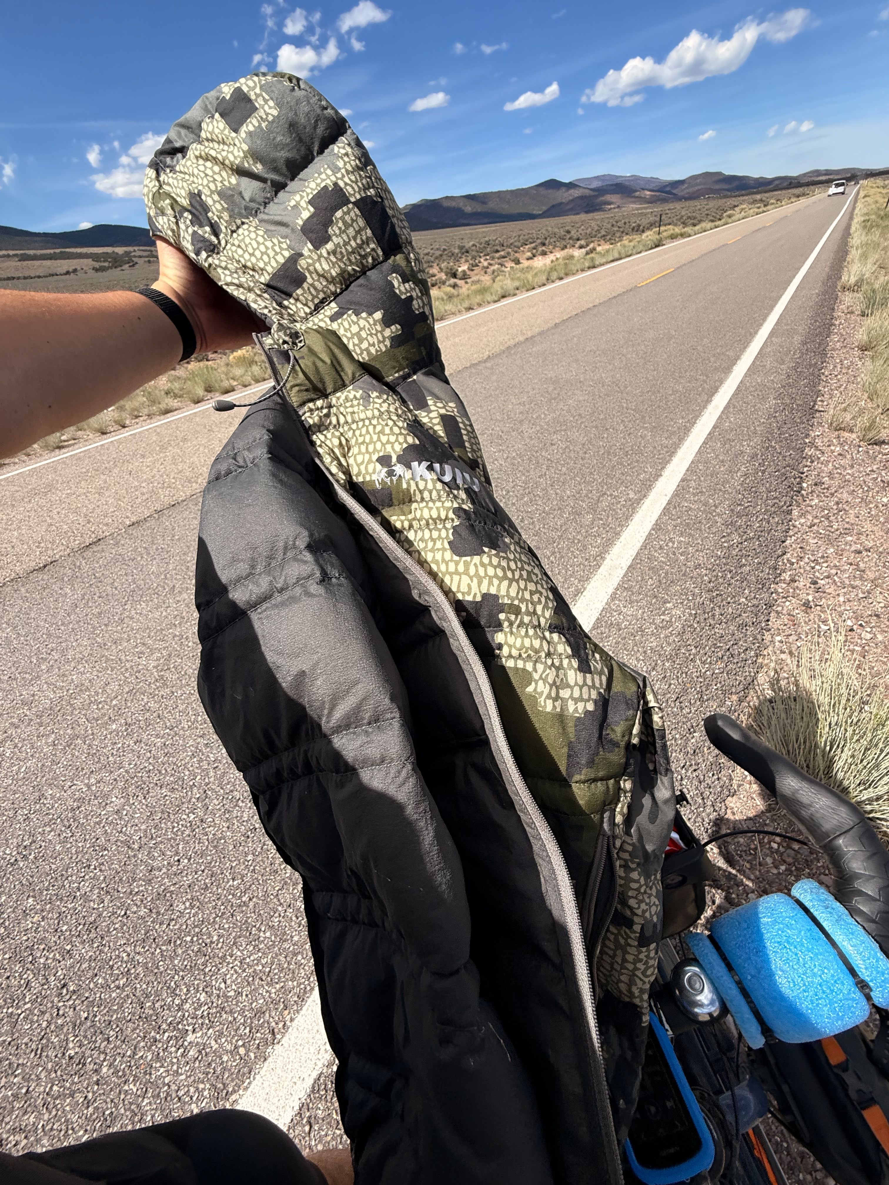 A camouflage Kuiu jacket held up on an empty road, the bicycle in the foreground