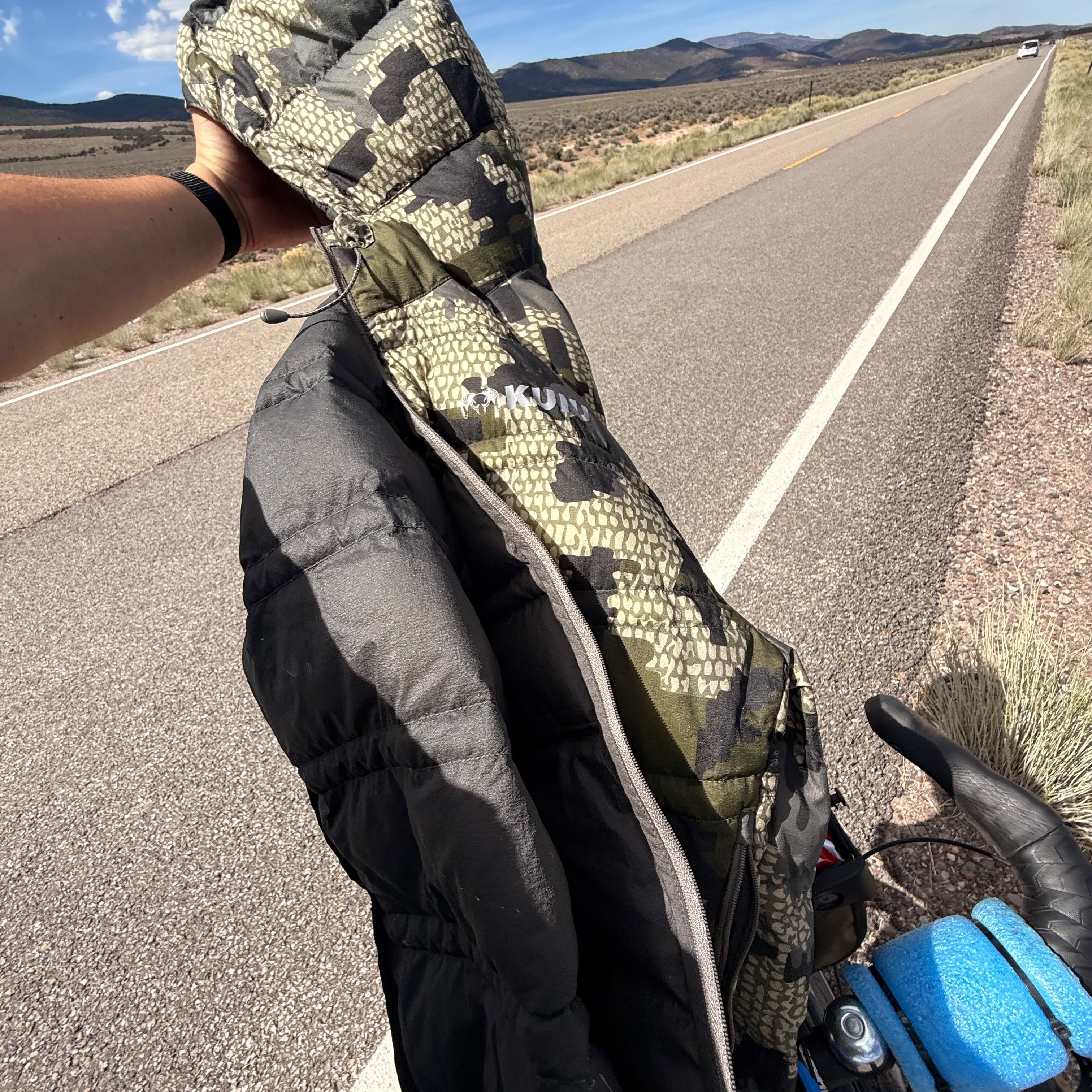 A camouflage Kuiu jacket held up on an empty road, the bicycle in the foreground