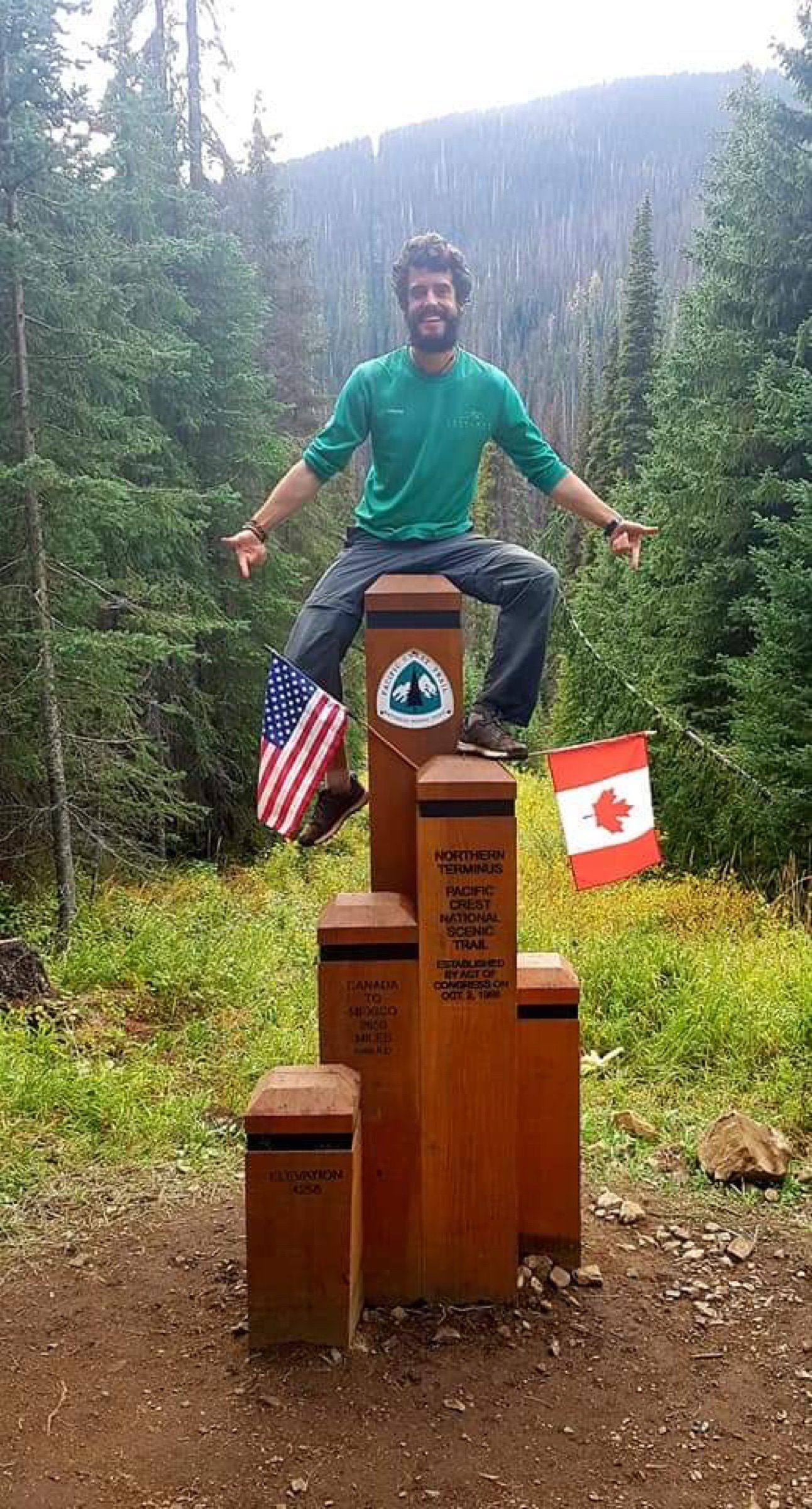 A bearded man sitting on the wooden northern terminus marker of the Pacific Crest Trail, between a US and a Canadian flag.